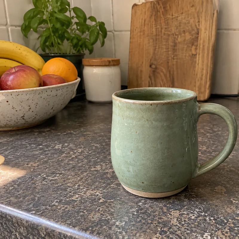A sage-green ceramic mug on a speckled kitchen counter, next to a fruit bowl.